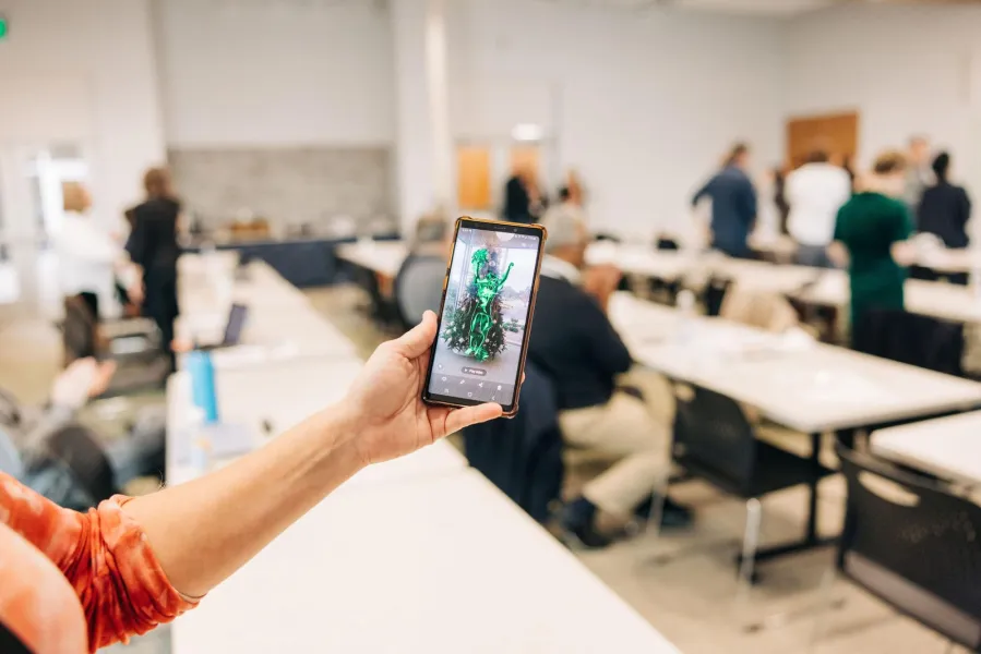 An arm extended out, holding a phone up, on the screen appears to be an AR sculpture.