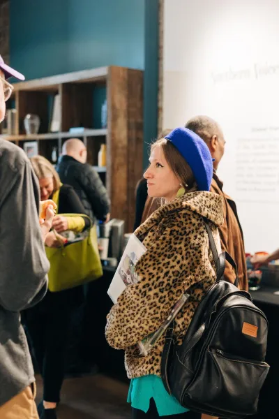 Woman in a leopard print jacket and purple beret standing in a room of other people, listening to someone.