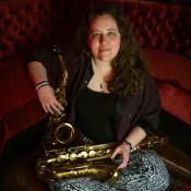 Woman sitting on a red rug in front of a velvet couch. She is holding two saxophones and smiling. 