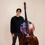 Man wearing black and grey, standing with an upright bass that has lights installed on parts of it.