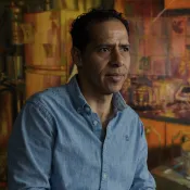 Person sitting in front of a warm, abstract painting in a collared blue shirt.