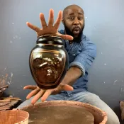 Person sitting at a pottery wheel holding out a black pot with a gold face.