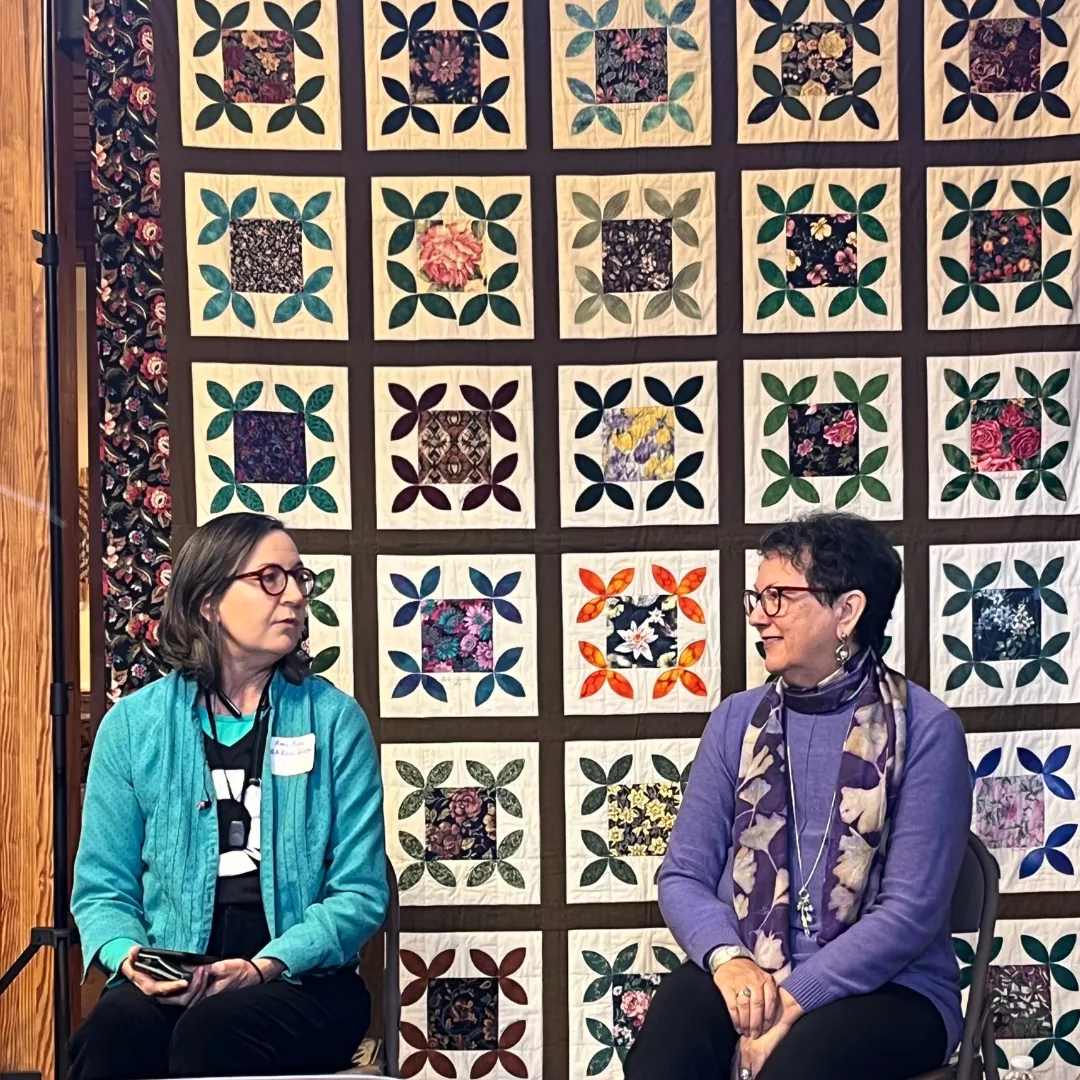 Two women sitting, facing each other, in front of a quilt hung on the wall